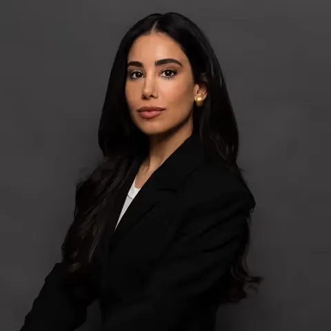 Portrait of a confident woman with long dark hair wearing a black blazer and gold earrings against a dark background.